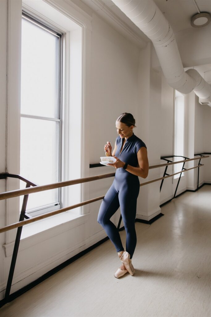 Lili Pfeifer of The Barbell Ballerina eating from a bowl while standing at the barre in navy unitard during nutrition coaching brand photography Minneapolis session
