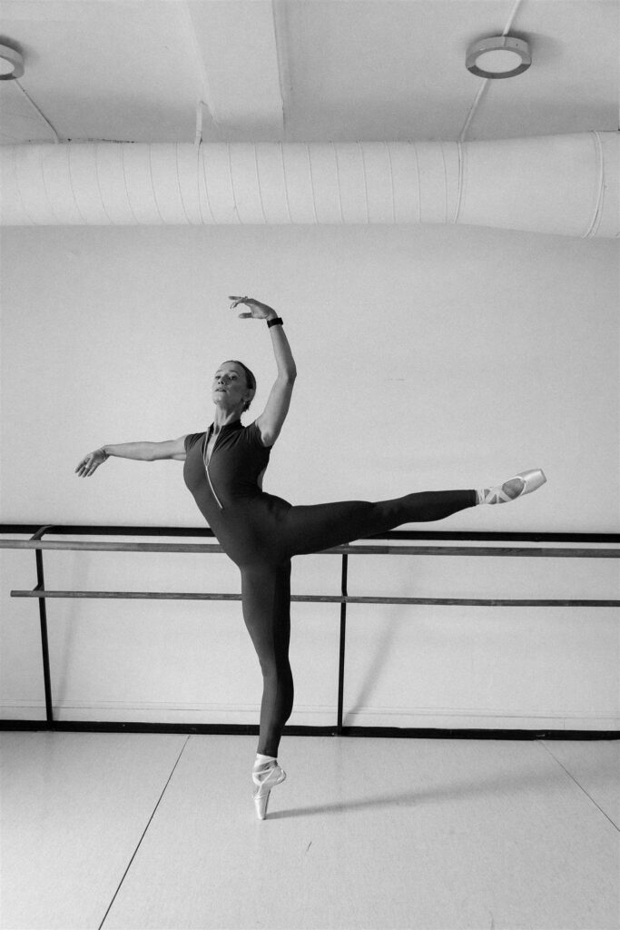 Lili Pfeifer of The Barbell Ballerina in black and white performing an arabesque at the barre in dance studio during fitness brand photography Minneapolis session