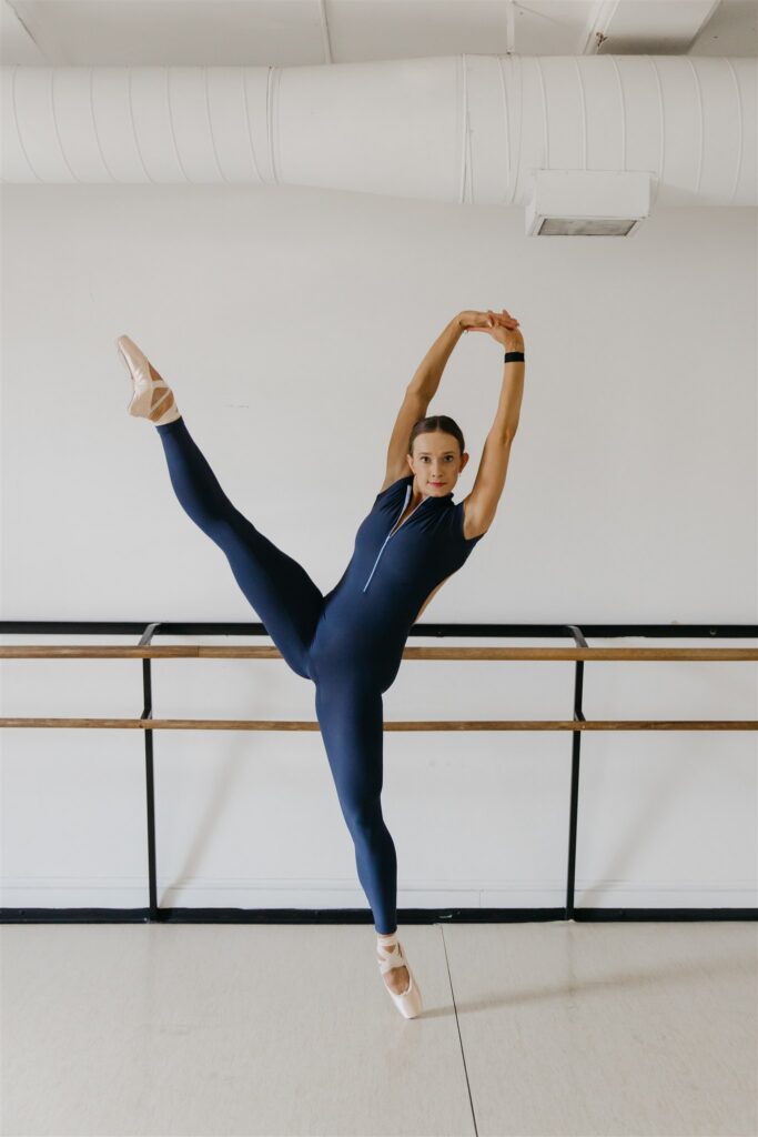 Lili Pfeifer of The Barbell Ballerina performing a grand battement facing forward in navy unitard at the barre during fitness brand photography Minneapolis session by Mycah Bain Photography