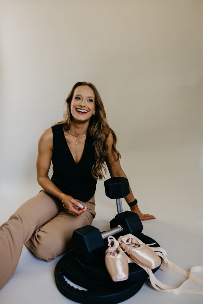 Lili Pfeifer of The Barbell Ballerina seated on floor laughing with dumbbells and pointe shoes during fitness brand photography Minneapolis session