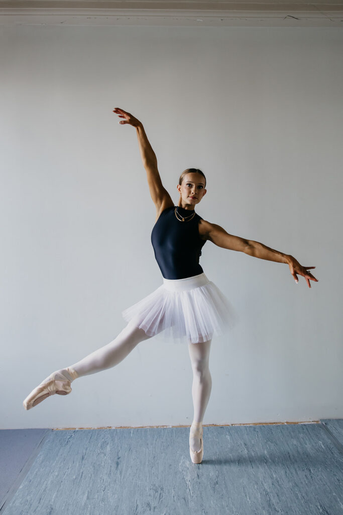 Lili Pfeifer of The Barbell Ballerina performing an attitude derrière in navy leotard and white tutu in dance studio during fitness brand photography Minneapolis session