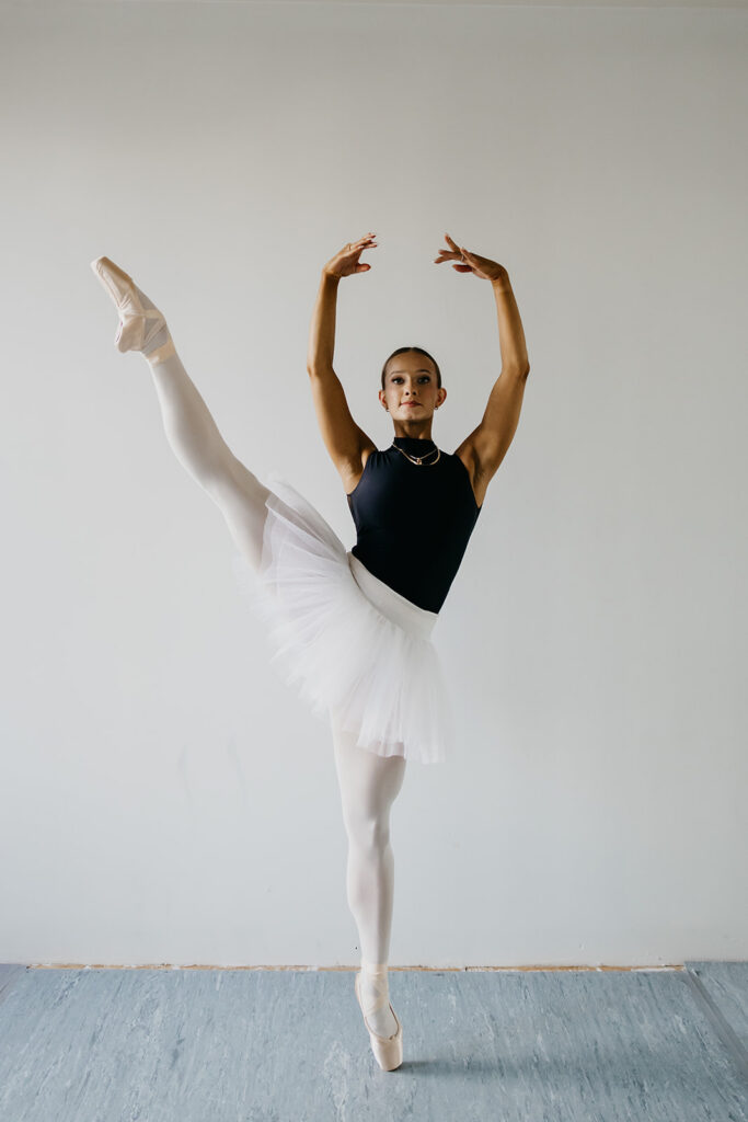 Lili Pfeifer of The Barbell Ballerina performing a high leg extension in pointe shoes and white tutu during fitness brand photography Minneapolis session by Mycah Bain Photography