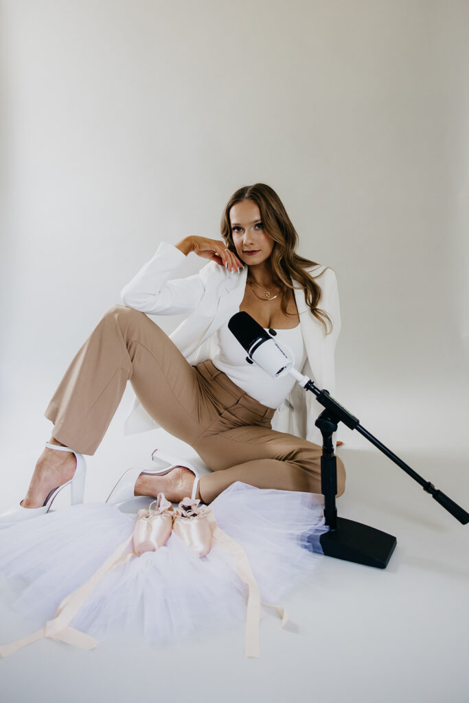 Lili Pfeifer of The Barbell Ballerina seated on floor in white blazer and tan trousers with microphone and pointe shoes during fitness brand photography Minneapolis session