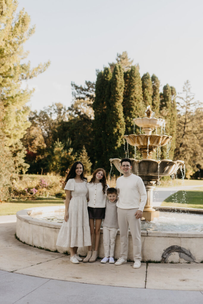 Family of four standing together beside an ornate stone fountain in coordinated neutral outfits