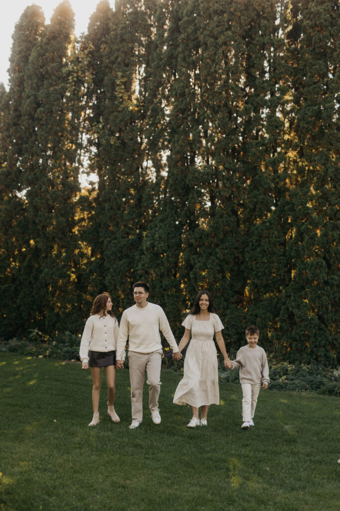 Family of four in neutral tones family photos outfits walking hand in hand across a lawn with tall evergreen trees behind them