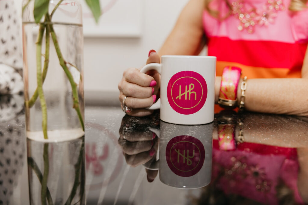 Close-up of a hand holding a white Haute House Studio branded mug on a reflective black table with glass bud vases in the background
