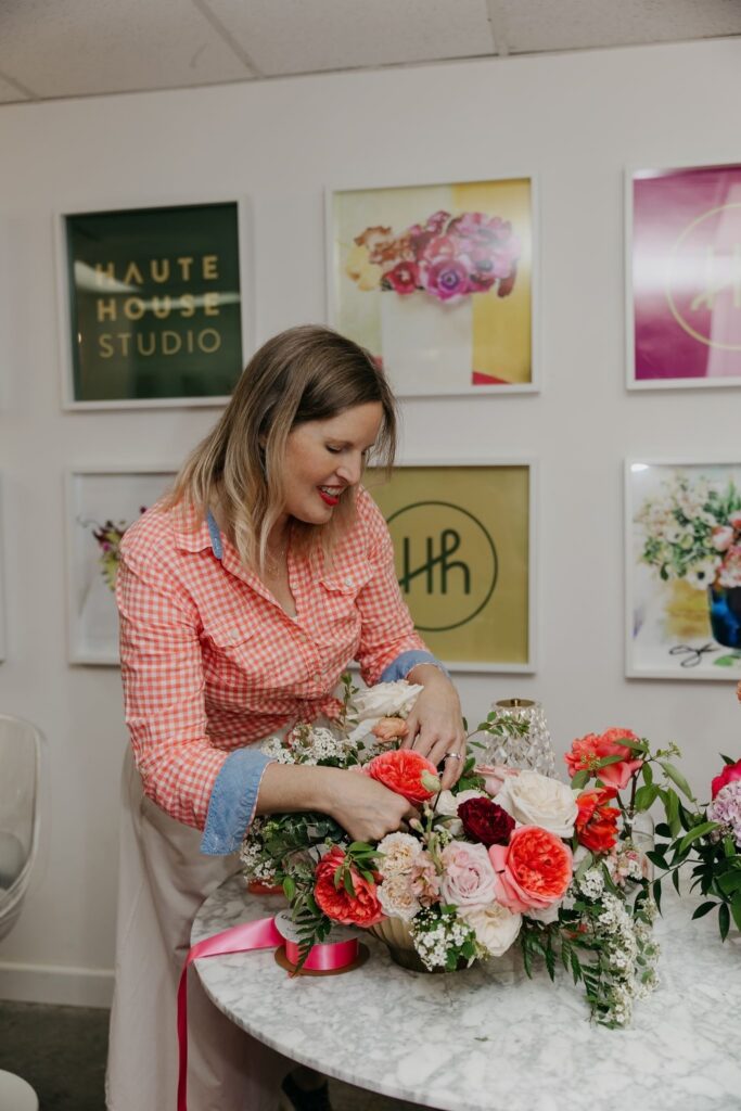 Haute House Studio team member in a pink gingham top arranging a lush floral centerpiece in front of the studio gallery wall Edina MN