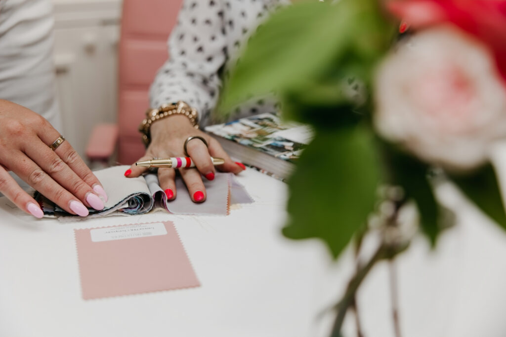 Close-up of two sets of hands reviewing fabric swatches and inspiration photos at a white table with a blush rose in the foreground during Haute House Studio brand photography session