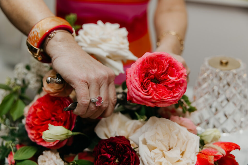 Close-up overhead shot of hands arranging red ranunculus coral roses and greenery into a floral centerpiece during Haute House Studio brand session