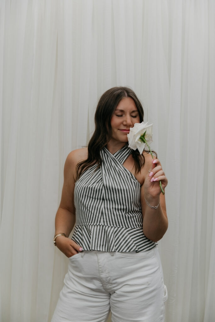 Haute House Studio team member in a striped halter top smelling a white garden rose against a white backdrop during brand session Minneapolis