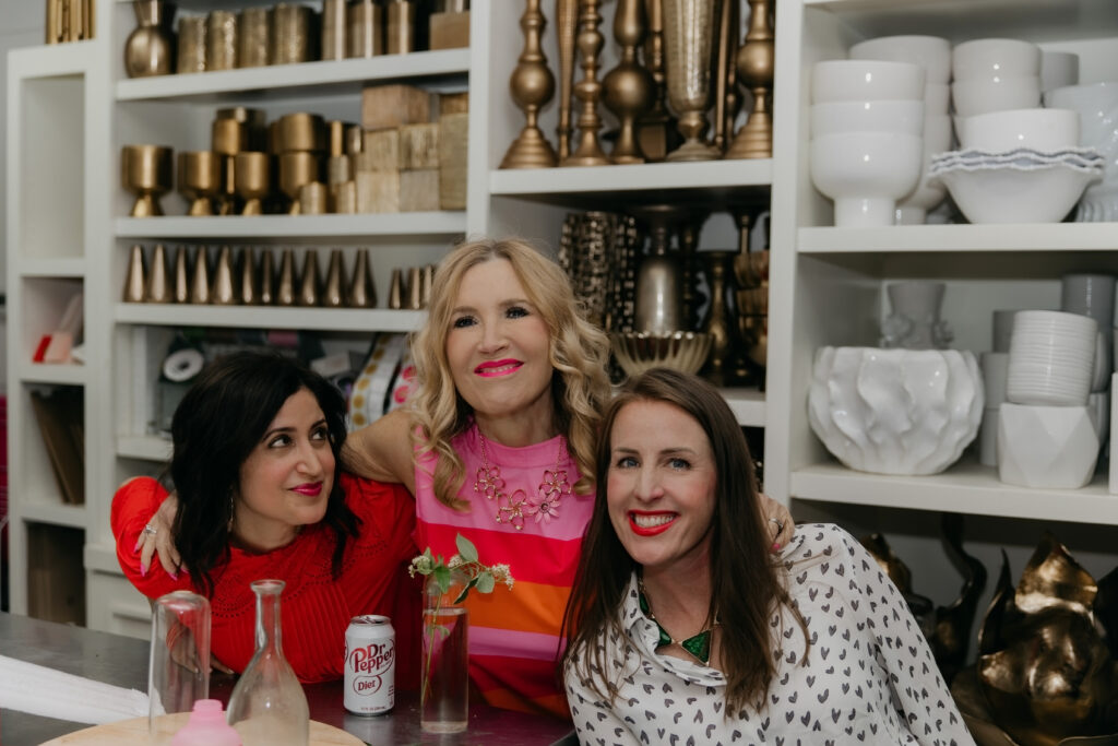 Three Haute House Studio team members smiling together in front of a shelving display of vases and event supplies in their Edina studio