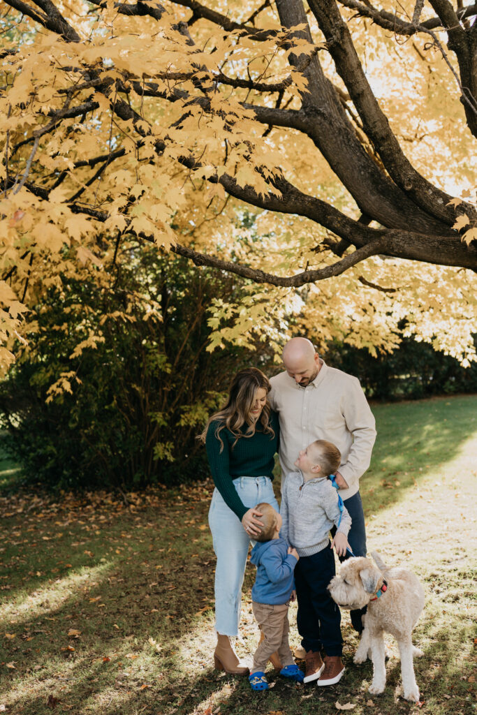 outdoor family photoshoot idea of parents standing with their two sons and dog under a golden fall tree