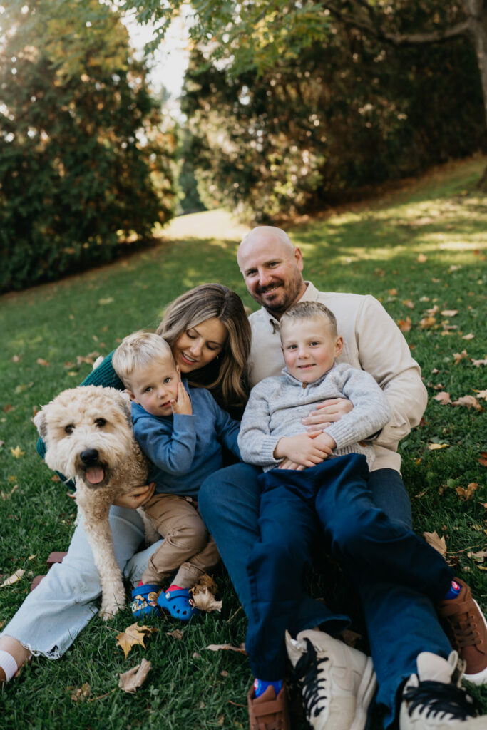 outdoor family photoshoot idea of parents and two sons sitting together on the grass with their dog leaning in