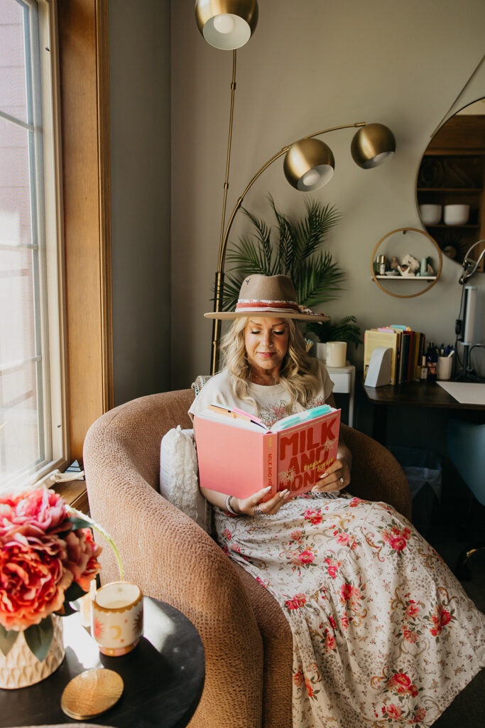 Women's wellness coach reading a book in a velvet armchair surrounded by flowers and gold lamps during brand photography session in Minneapolis