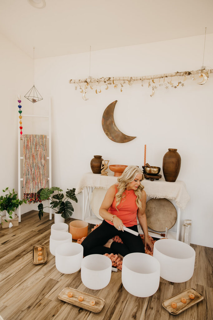 Women's energy healer seated on the floor surrounded by crystal singing bowls in a bohemian sound healing studio during brand photography session