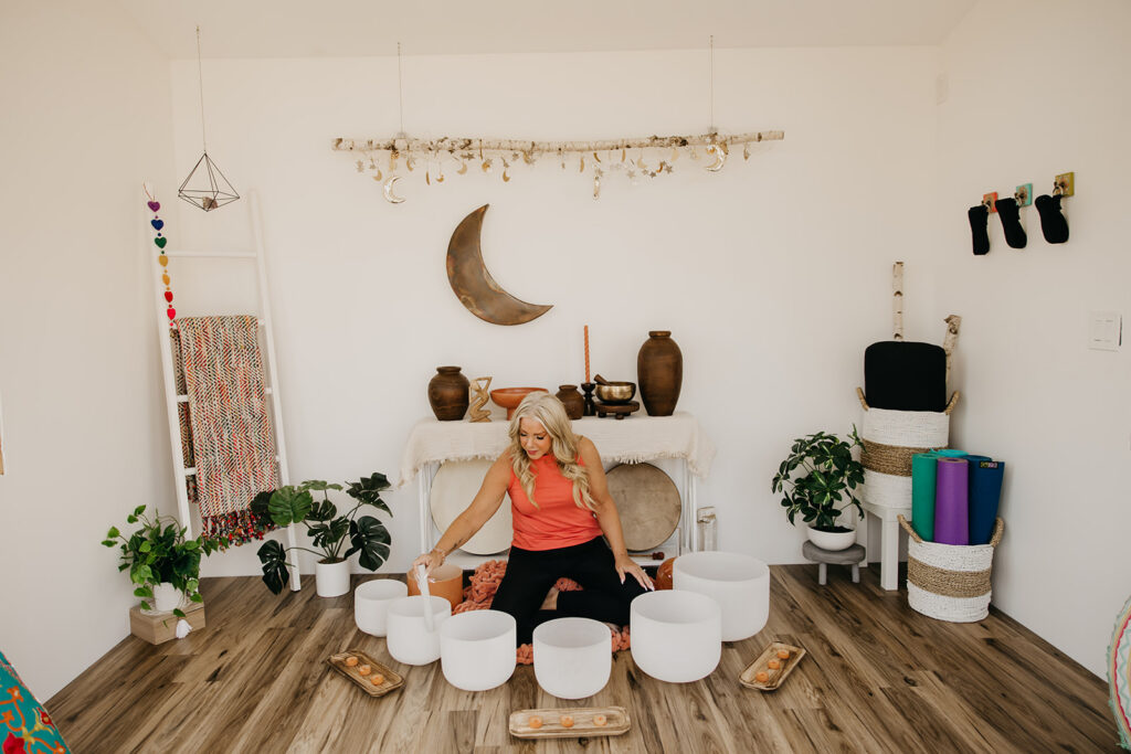 Women's wellness coach seated with singing bowls in front of white altar with crescent moon decor during brand photography session