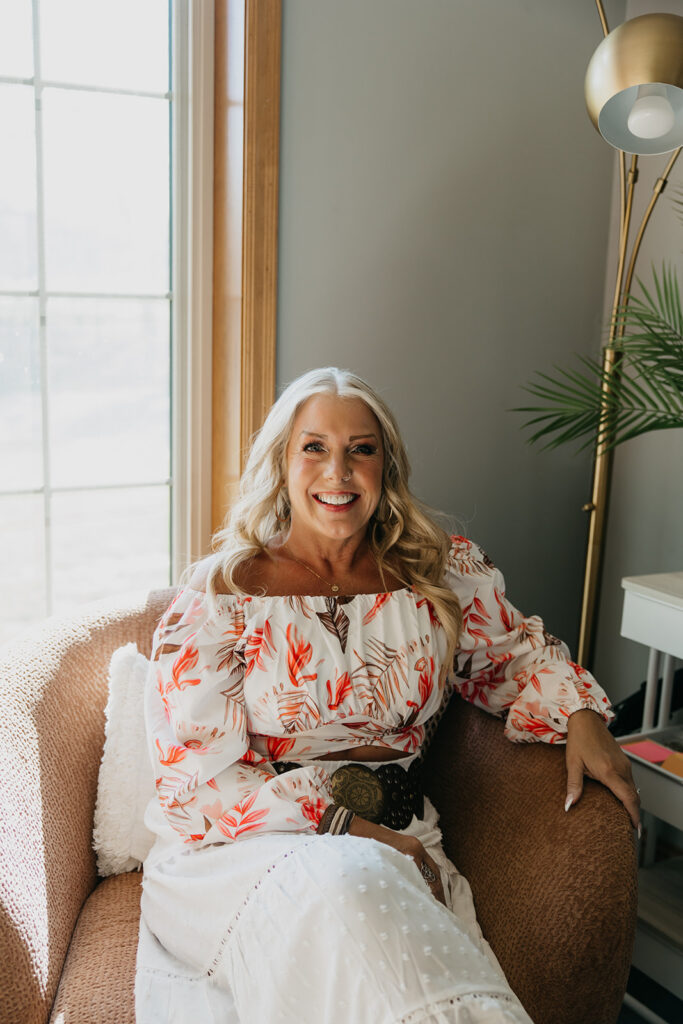 Women's wellness coach smiling while seated in a cozy chair by a sunny window during brand photography session in Minneapolis