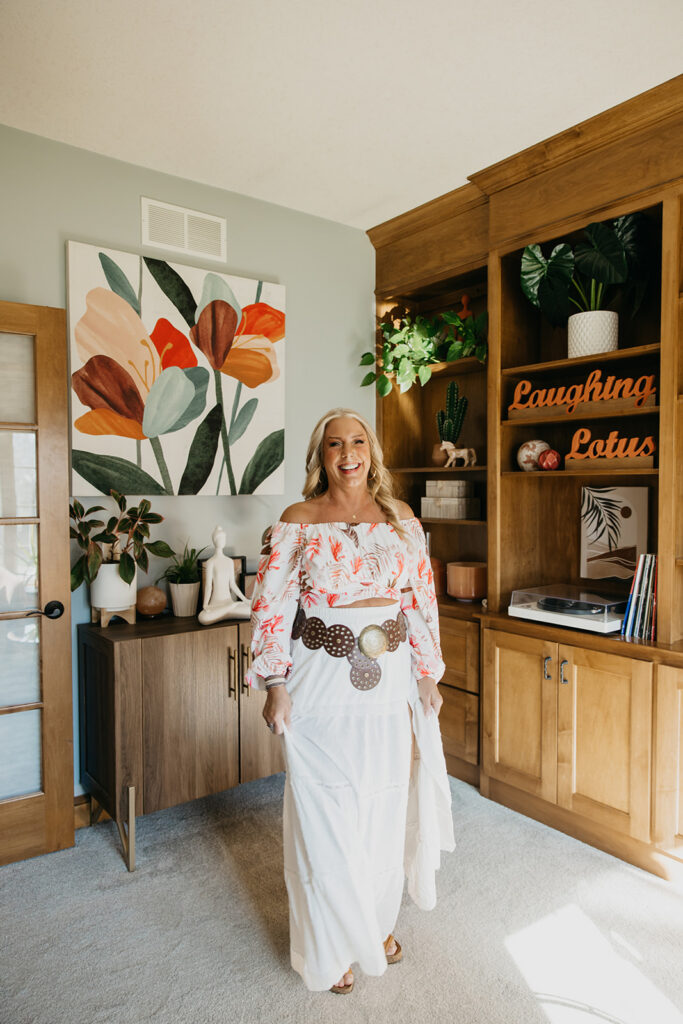 Full-length portrait of women's wellness coach in white skirt and floral top standing in front of Laughing Lotus home studio bookcase