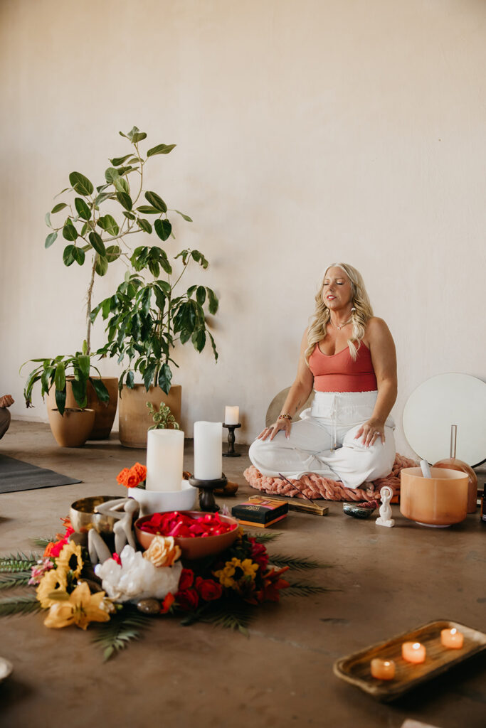 Women's wellness coach seated in meditation pose surrounded by candles, flowers, and plants in a retreat-style space during brand photography session