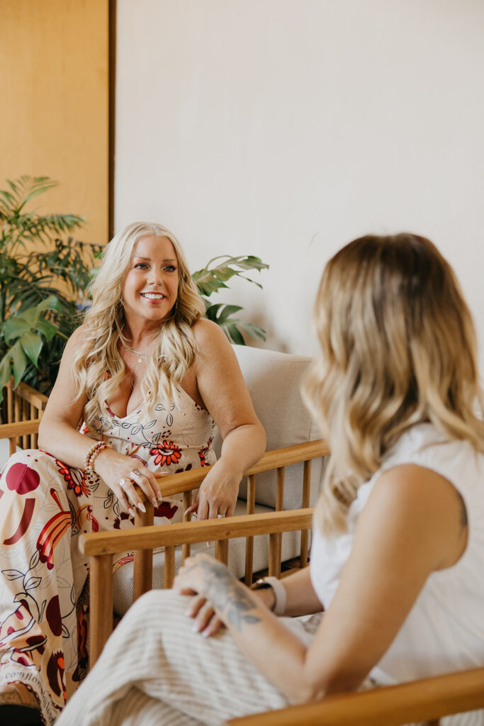 Women's wellness coach and photographer in conversation during brand photo shoot, seated across from each other in a bright open space