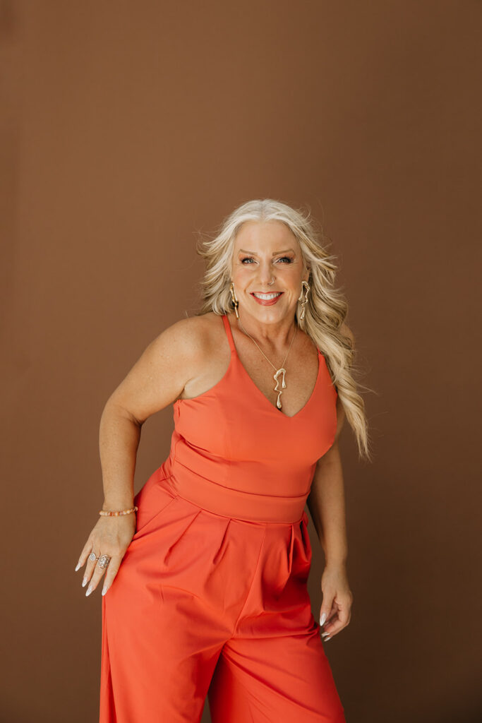 Women's wellness coach leaning forward and smiling in a bright orange jumpsuit during brand photography session on brown backdrop
