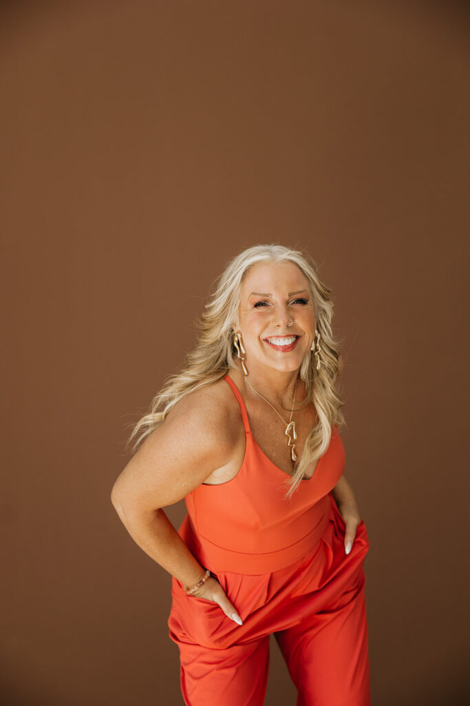 Women's wellness coach posing with hand on hip in an orange jumpsuit against a brown studio backdrop during brand shoot