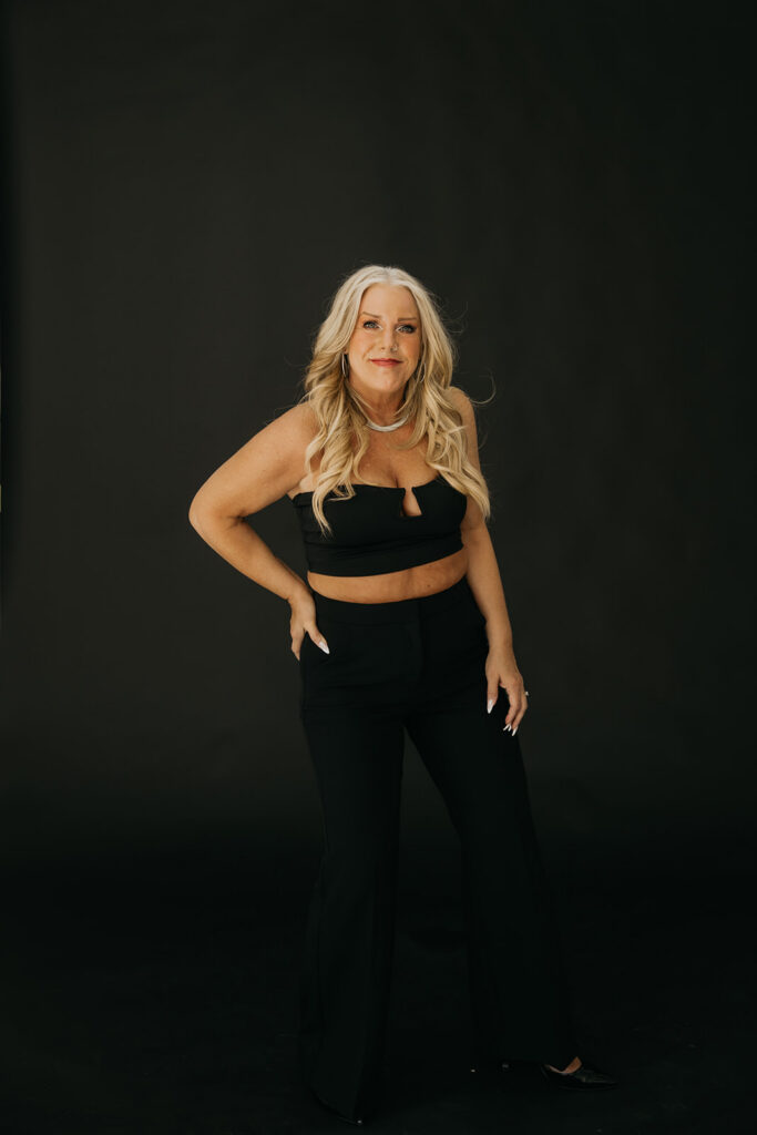 Women's mentor standing with hands on hips in all-black outfit during Minneapolis brand photo shoot on dark backdrop