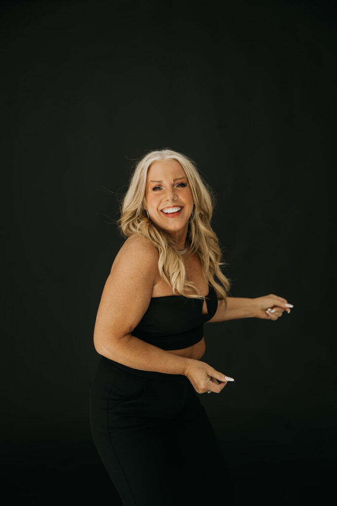 Women's wellness coach laughing and pointing during bold brand photography session on dark studio backdrop