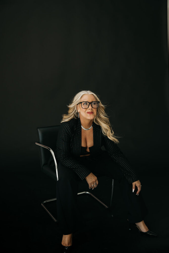 Women's wellness coach laughing while seated in a black chair wearing a black blazer and glasses during brand photography session