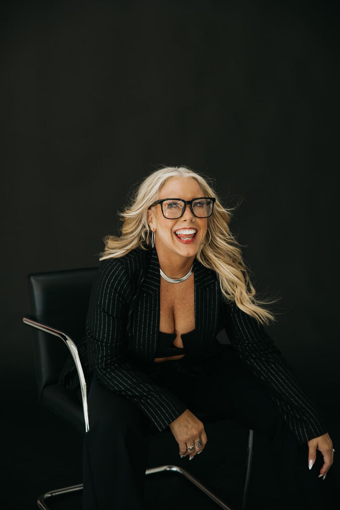 Women's mentor smiling with wind-blown hair in a black blazer during brand photo shoot on dark backdrop