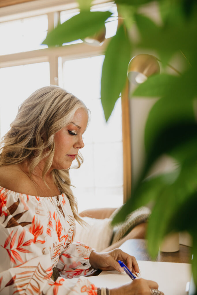 Close-up of women's wellness coach writing at her desk, shot through green plant leaves during brand photography session