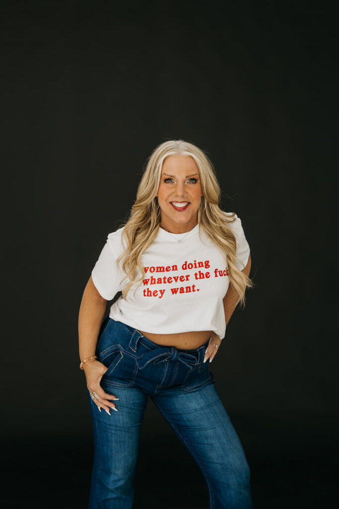 Women's mentor in graphic feminist tee with hands on hips laughing during brand photo shoot on dark backdrop