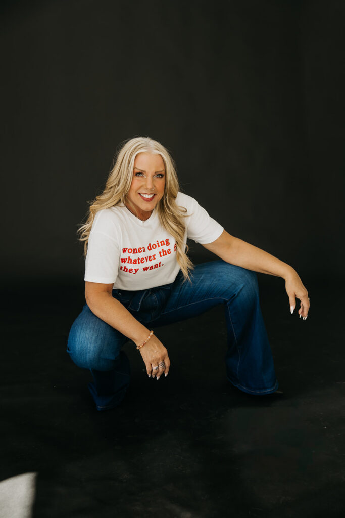 Women's mentor crouching in a playful pose in jeans and statement graphic tee during brand photo shoot on dark backdrop