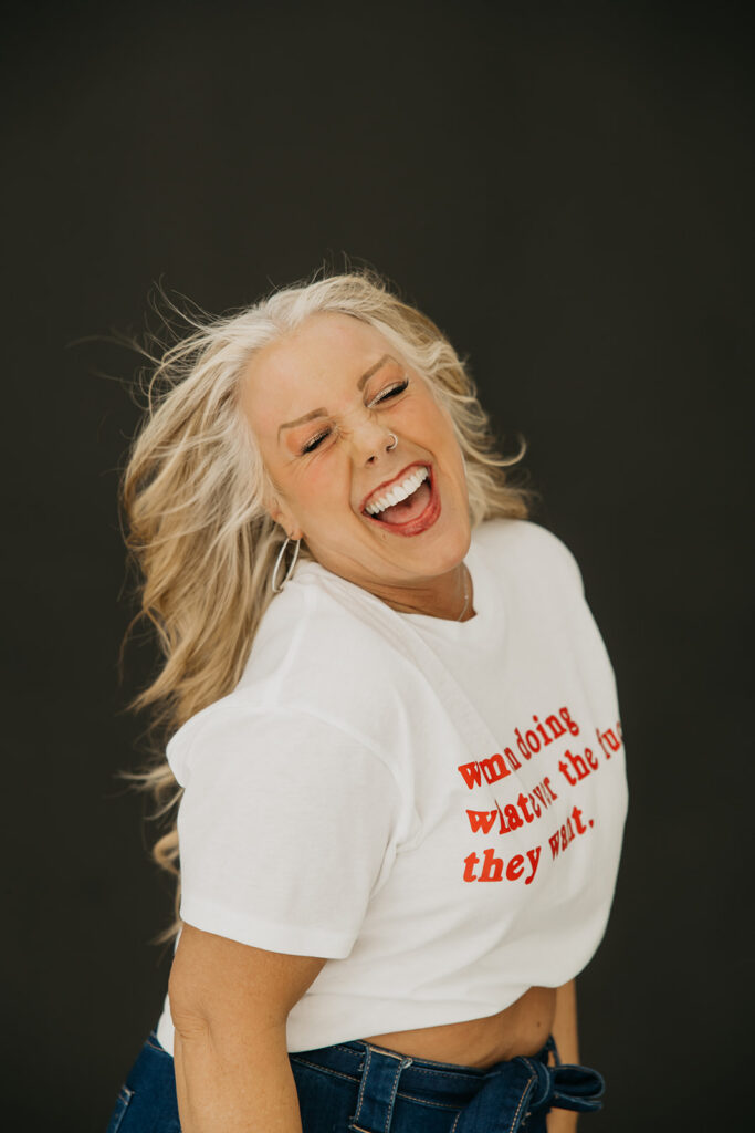 Women's wellness coach throwing her head back in full laughter with hair flying during brand photography session in Minneapolis, wearing a statement feminist graphic tee