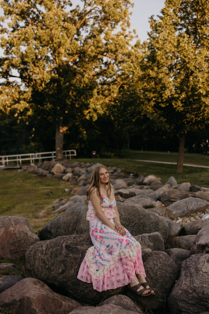 Hopkins High School senior girl in pink floral dress seated on boulder smiling at golden hour