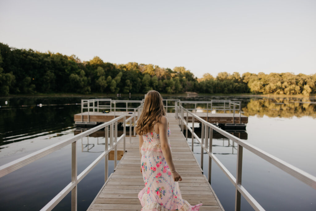 Hopkins High School senior girl in pink floral dress twirling on wooden dock with lake reflection behind her