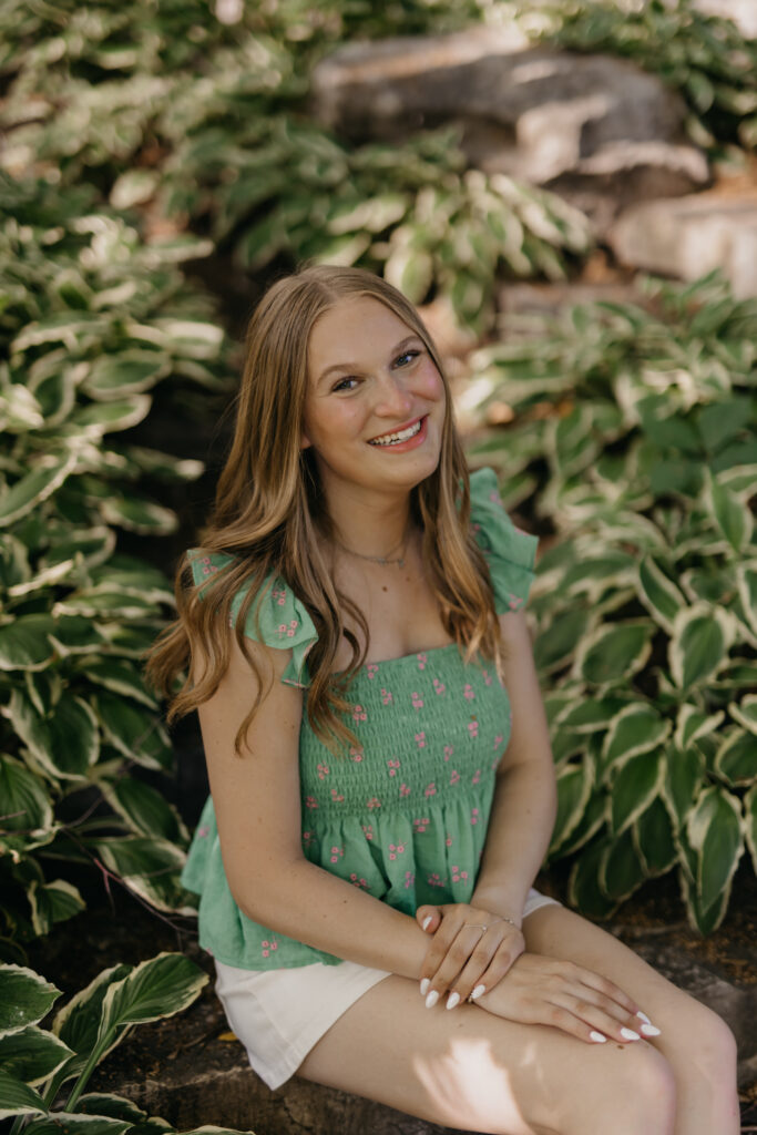 Hopkins High School senior girl in green top seated on rock surrounded by large leafy plants smiling