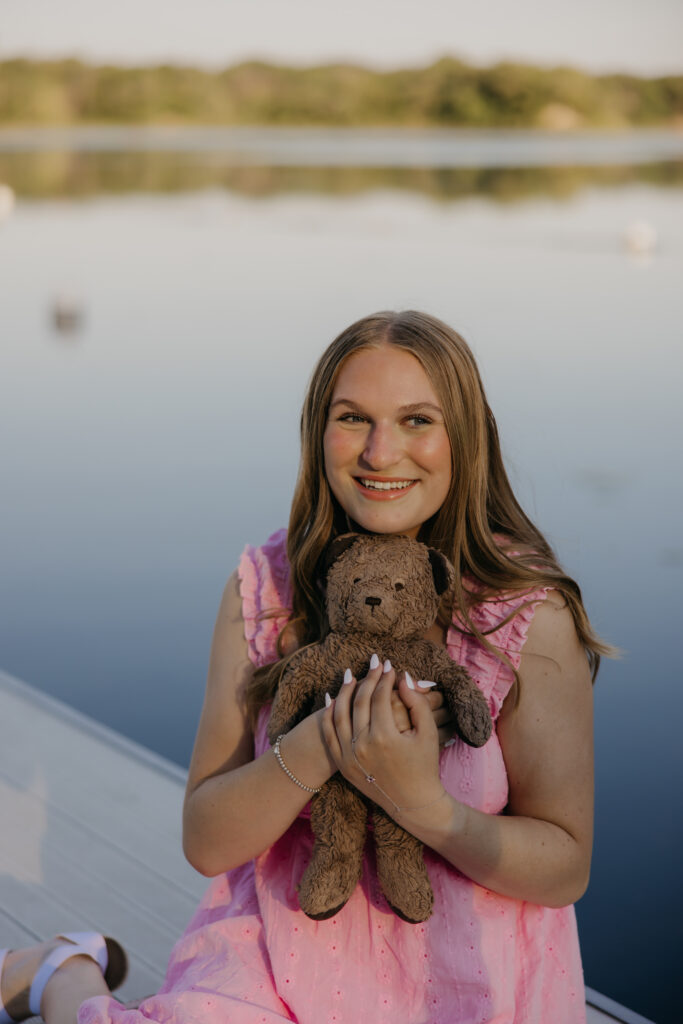 Hopkins High School senior girl in pink dress seated on dock holding stuffed bunny smiling at camera