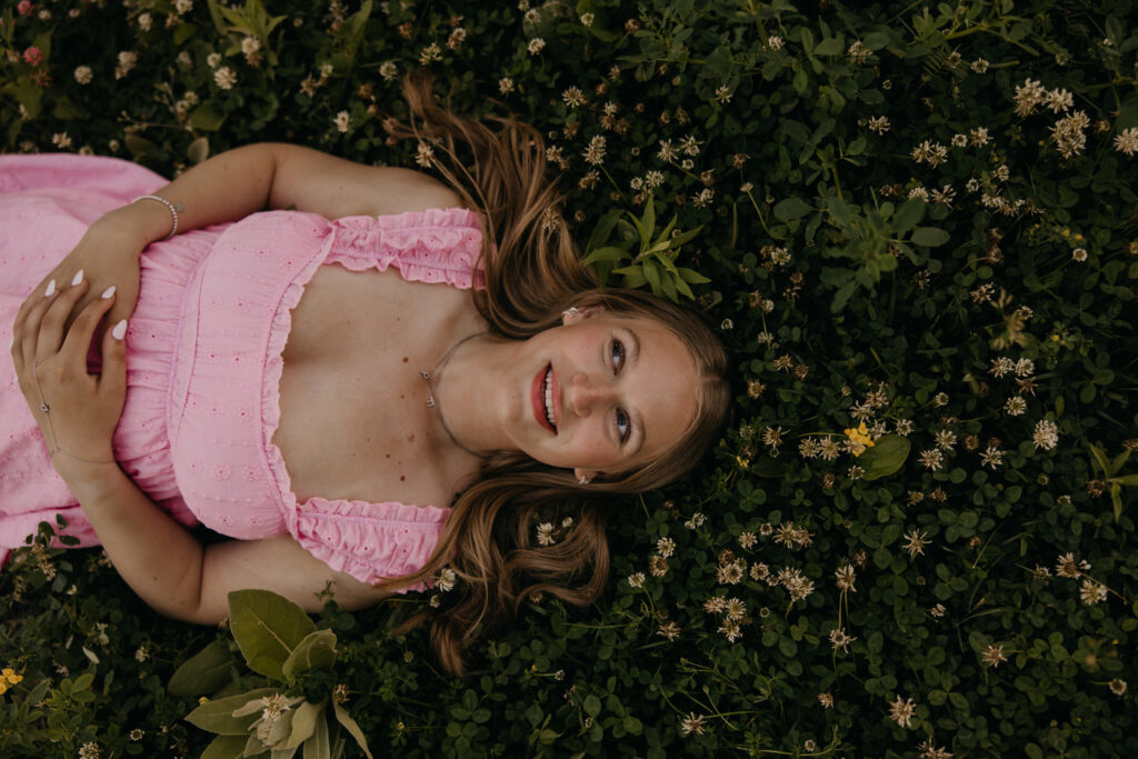 Hopkins senior photos close-up overhead of girl in pink dress lying in clover wildflower field smiling up at camera