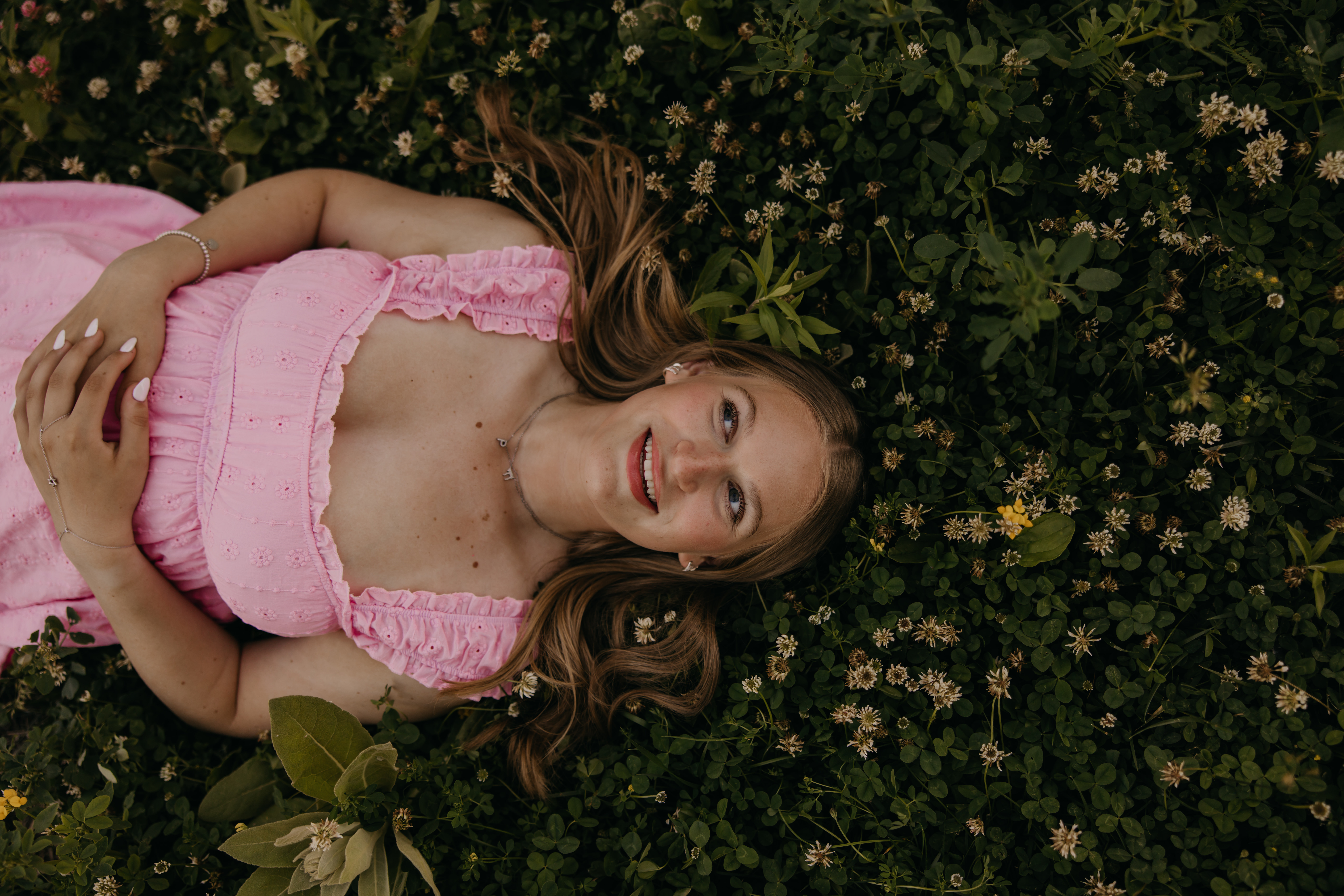 Hopkins senior photos of girl in pink eyelet dress lying in clover wildflower field smiling up at camera