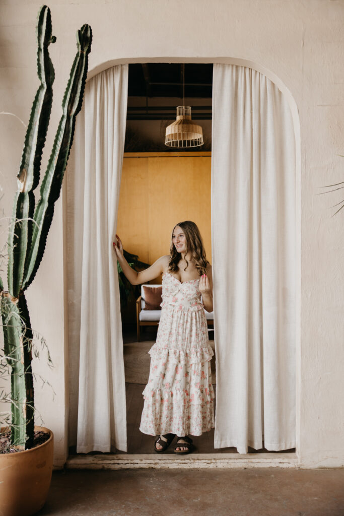 Hopkins High School senior girl pulling back white curtain in arched doorway with cactus beside her