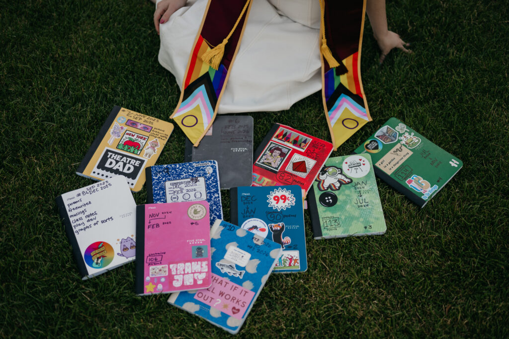 Overhead flat lay of hand-decorated journals fanned across grass, including "Theatre Dad" and "Trans Joy" covers, with graduation stole and dress hem visible