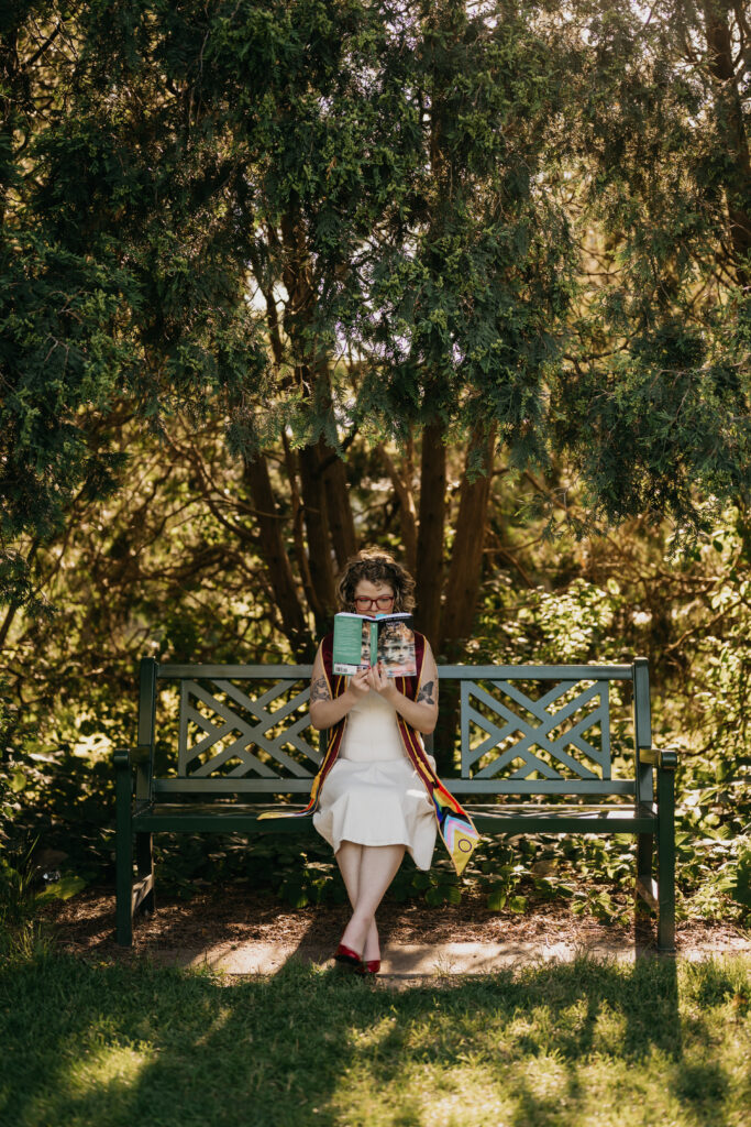 graduate seated on ornate garden bench reading a book held up in shaded light