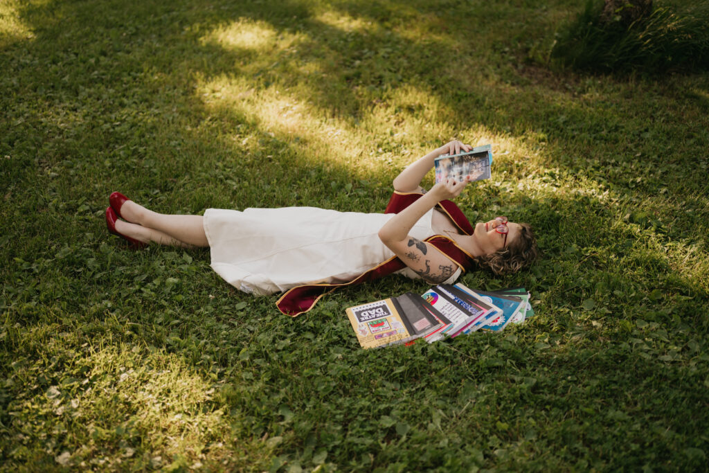 wide shot of graduate reclined in grass with journal raised, stole draped beside her