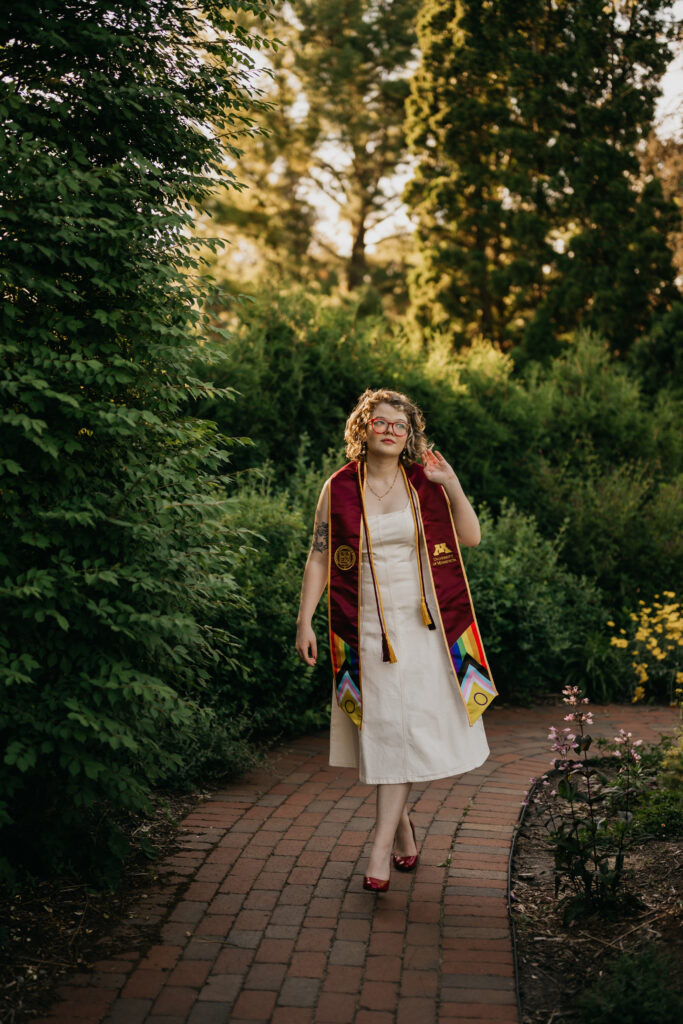 University of Minnesota graduate walking toward camera on a hedge-lined brick path