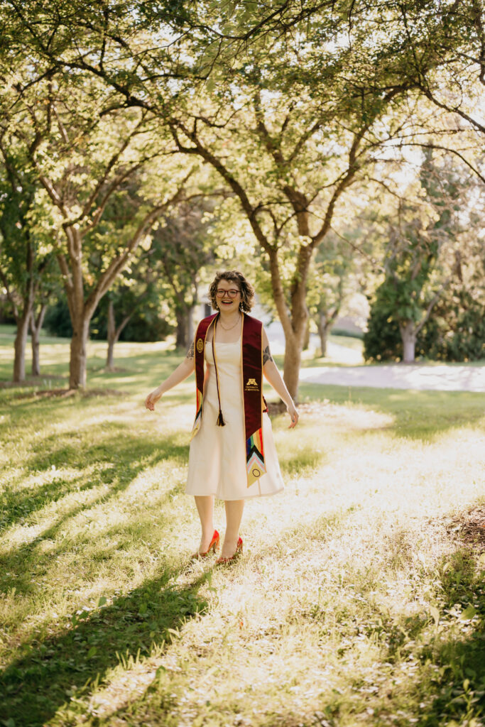 graduate laughing and walking under dappled tree light, arms open