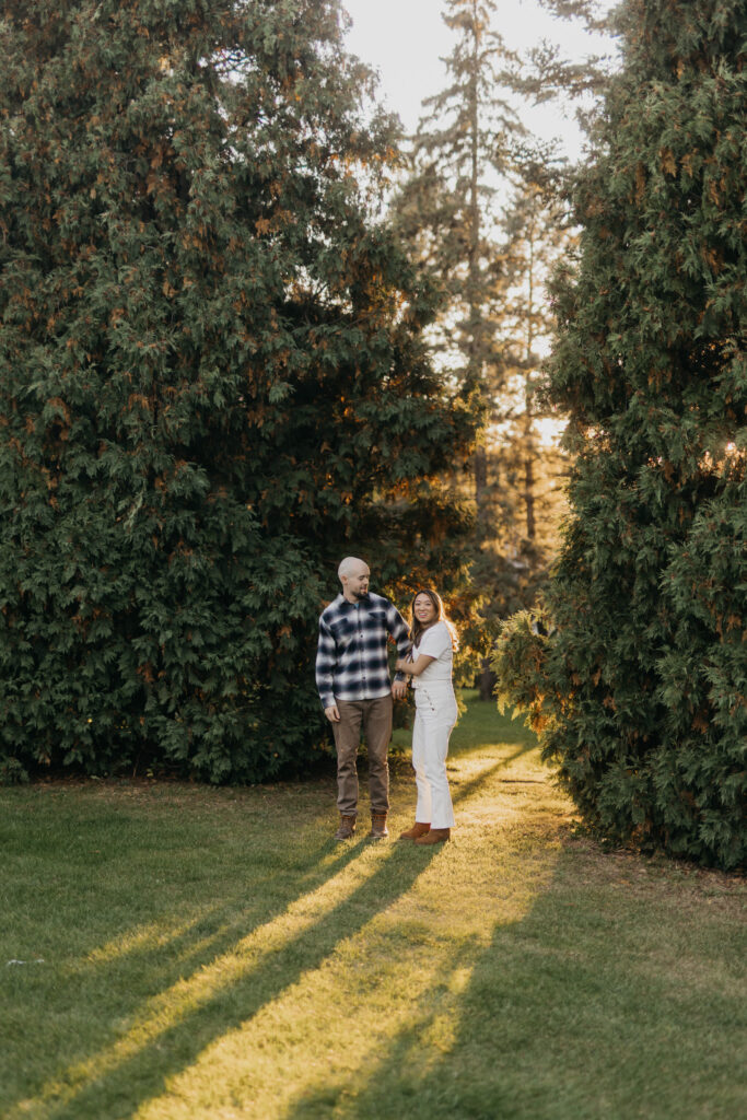 Couple standing together between tall evergreen trees, long shadows across the grass
