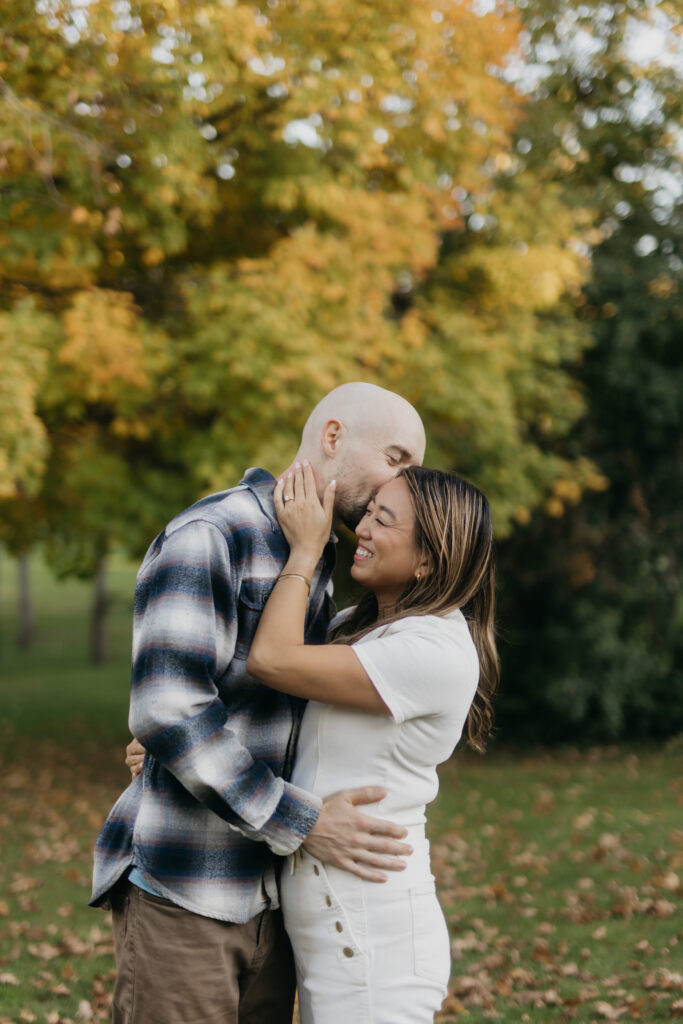 Couple embracing under golden fall foliage, man in plaid flannel kissing woman's forehead