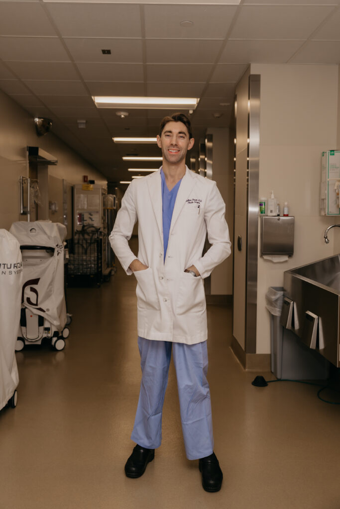 Dr. Matthew Braza in white coat and scrubs standing in hospital corridor near supply cart