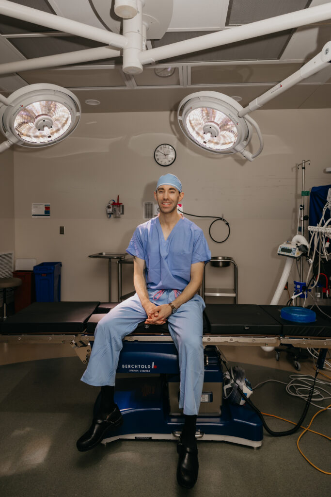 Dr. Matthew Braza in surgical cap and scrubs seated on operating table with hands clasped